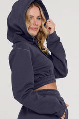 A woman wearing a navy blue cropped hoodie and jogger set, with the hoodie featuring a front pouch and the joggers having an elastic waistband. The woman is smiling and has her hood up, posing for the camera.