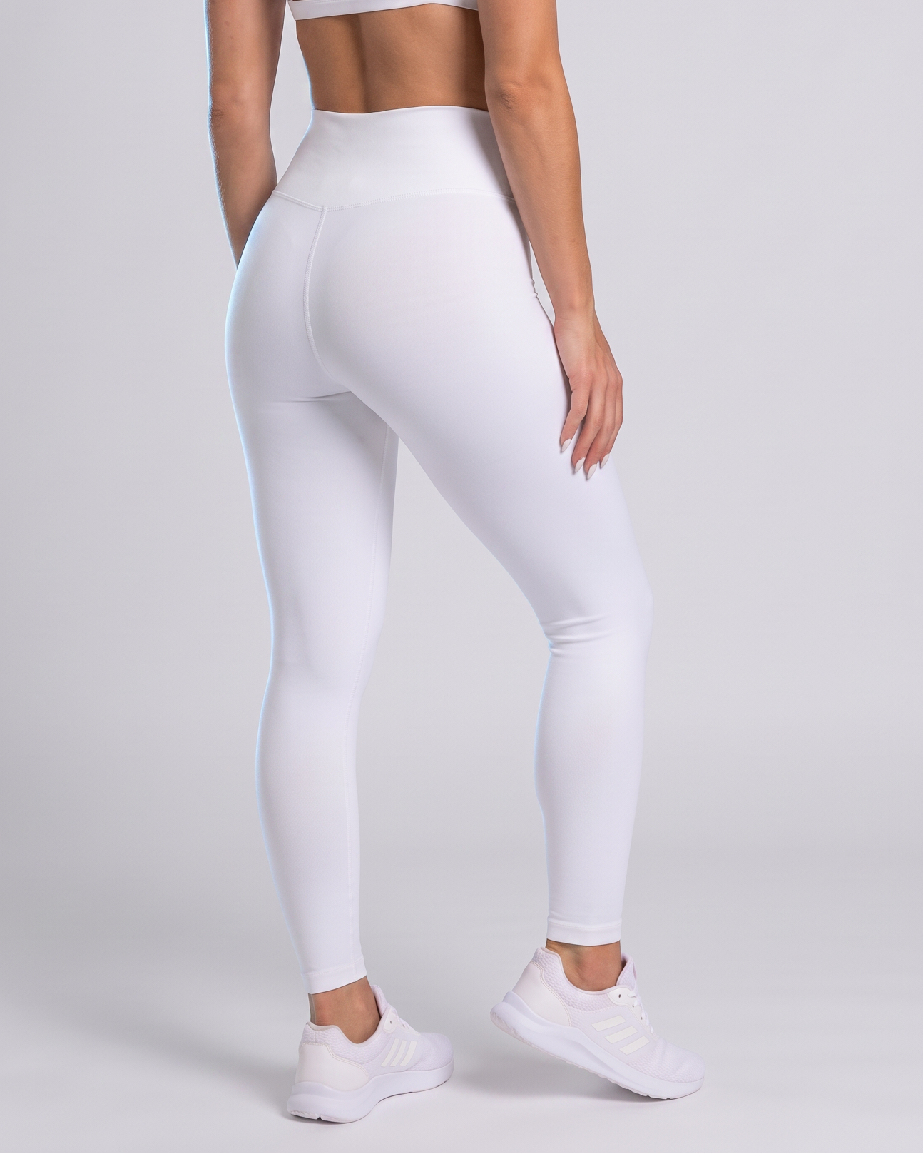 A woman shown from the back wearing a white sports bra with crisscross straps and high-waisted white leggings, standing against a light gray studio background.