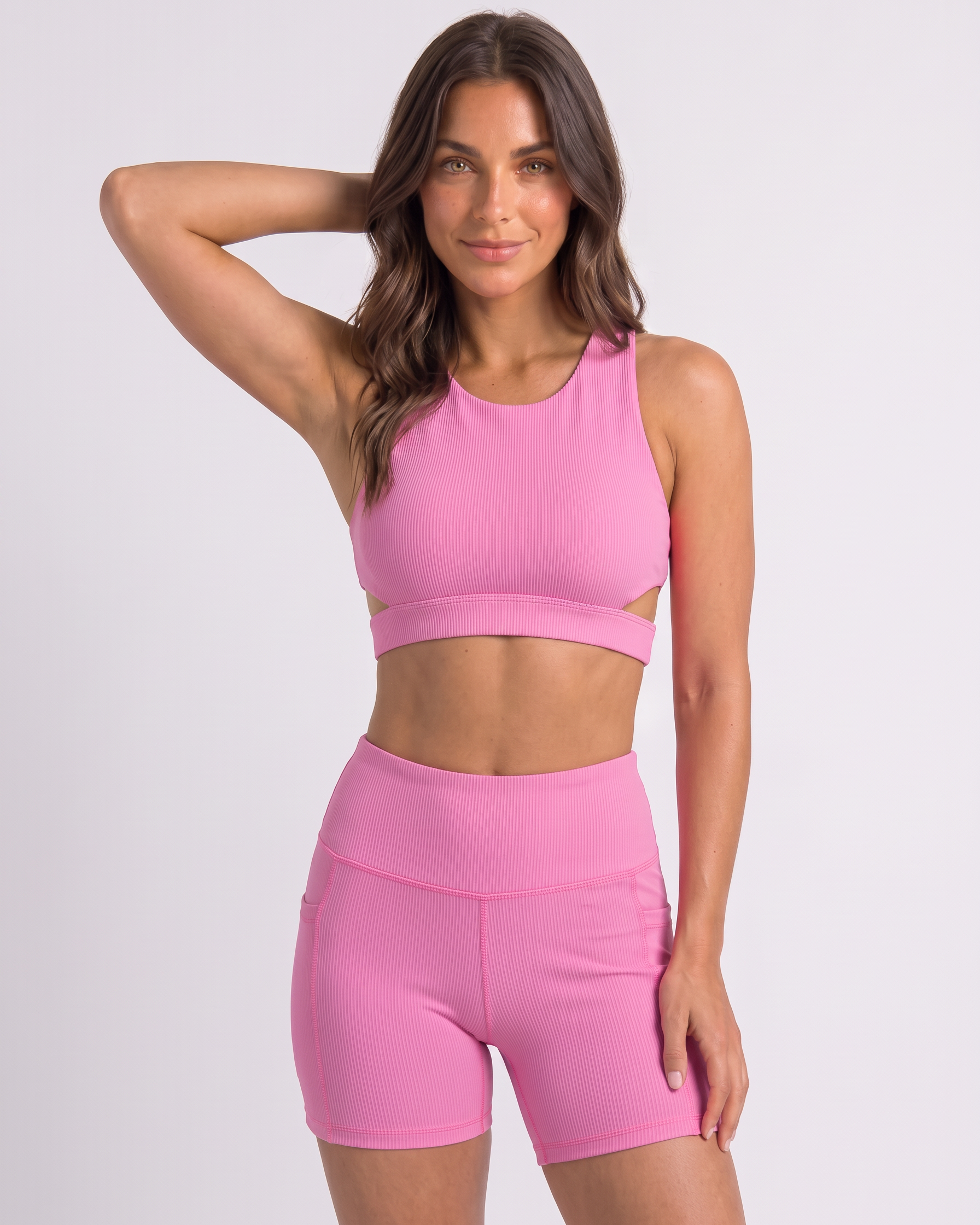 A woman modeling a matching pink activewear set consisting of a sports bra with side cutouts and high-waisted biker shorts, set against a plain light gray studio background.