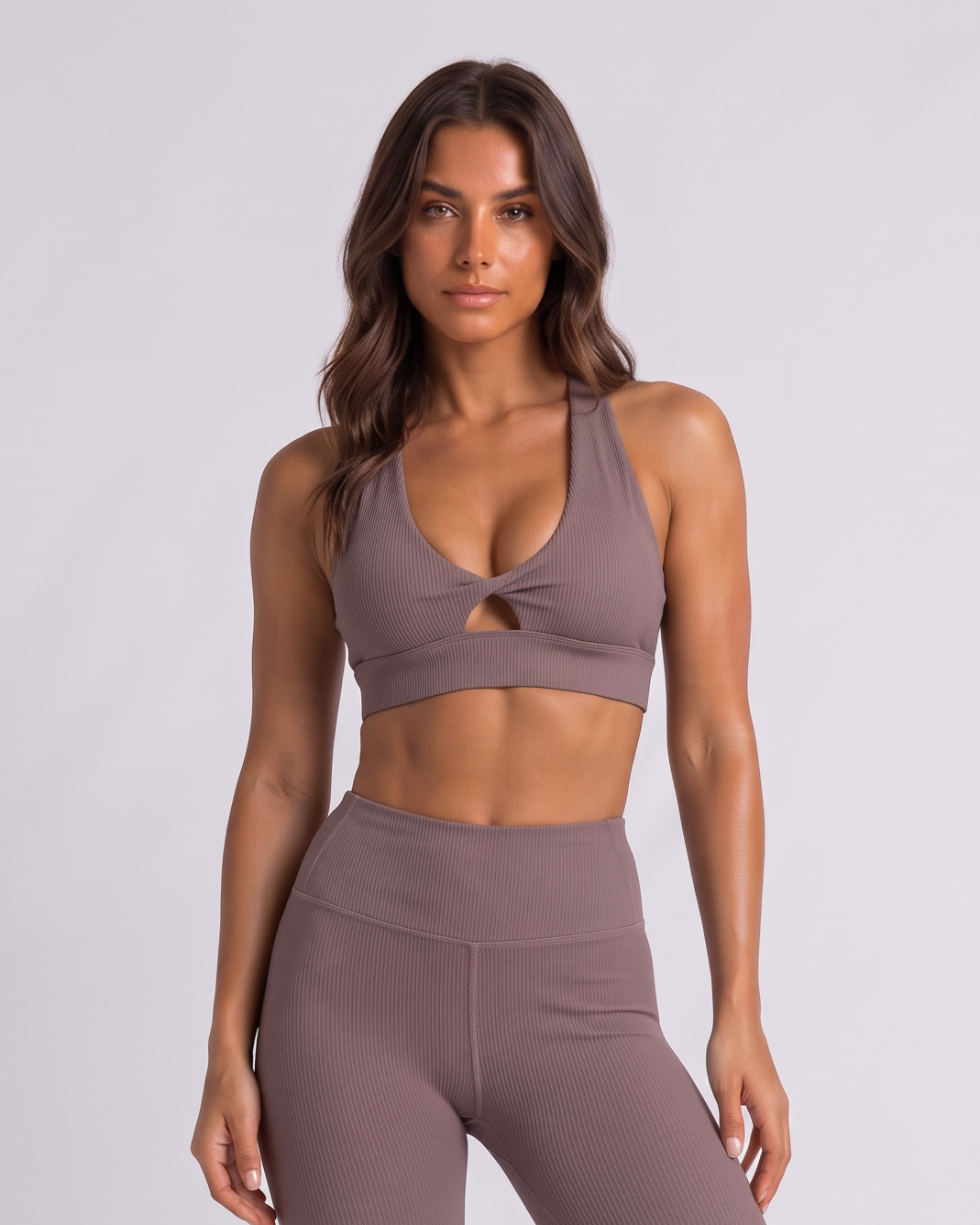 A woman modeling a matching two-piece activewear set consisting of a dusty mauve sports bra with a front keyhole cutout and high-waisted leggings, paired with white sneakers, against a light gray studio background.