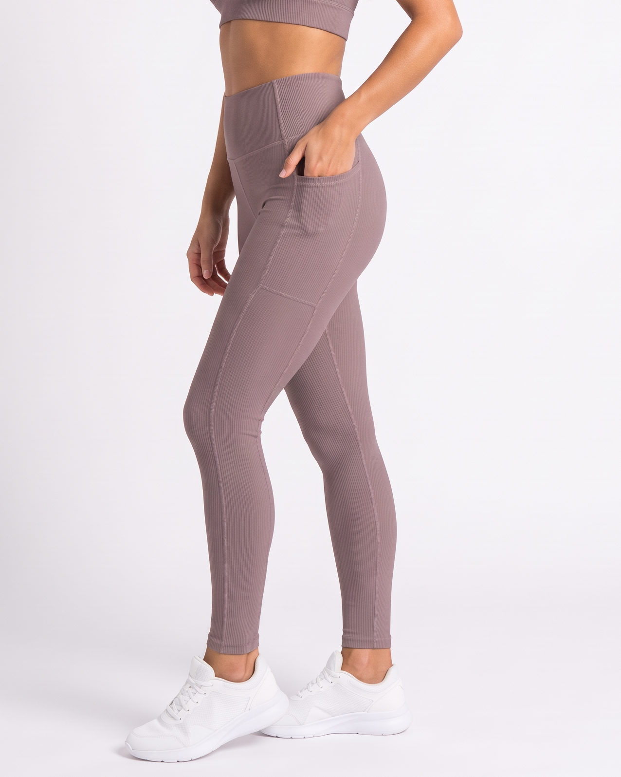 A woman modeling a matching dusty mauve activewear set consisting of a sports bra and high-waisted leggings with side pockets, paired with white sneakers, against a plain white studio background.