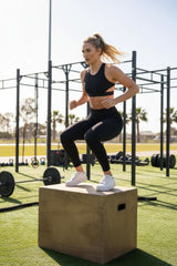 Woman performing box jumps in an outdoor fitness area with equipment and green grass.