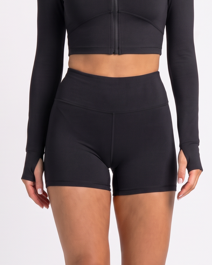 A woman modeling a black two-piece activewear set consisting of a long-sleeve mock-neck crop top with a front zipper and matching high-waisted shorts, paired with white sneakers, against a light gray studio background.