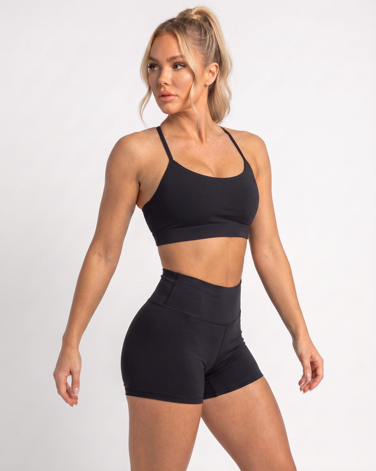 A woman modeling a black two-piece activewear set consisting of a sports bra and high-waisted shorts, photographed against a light gray studio background.