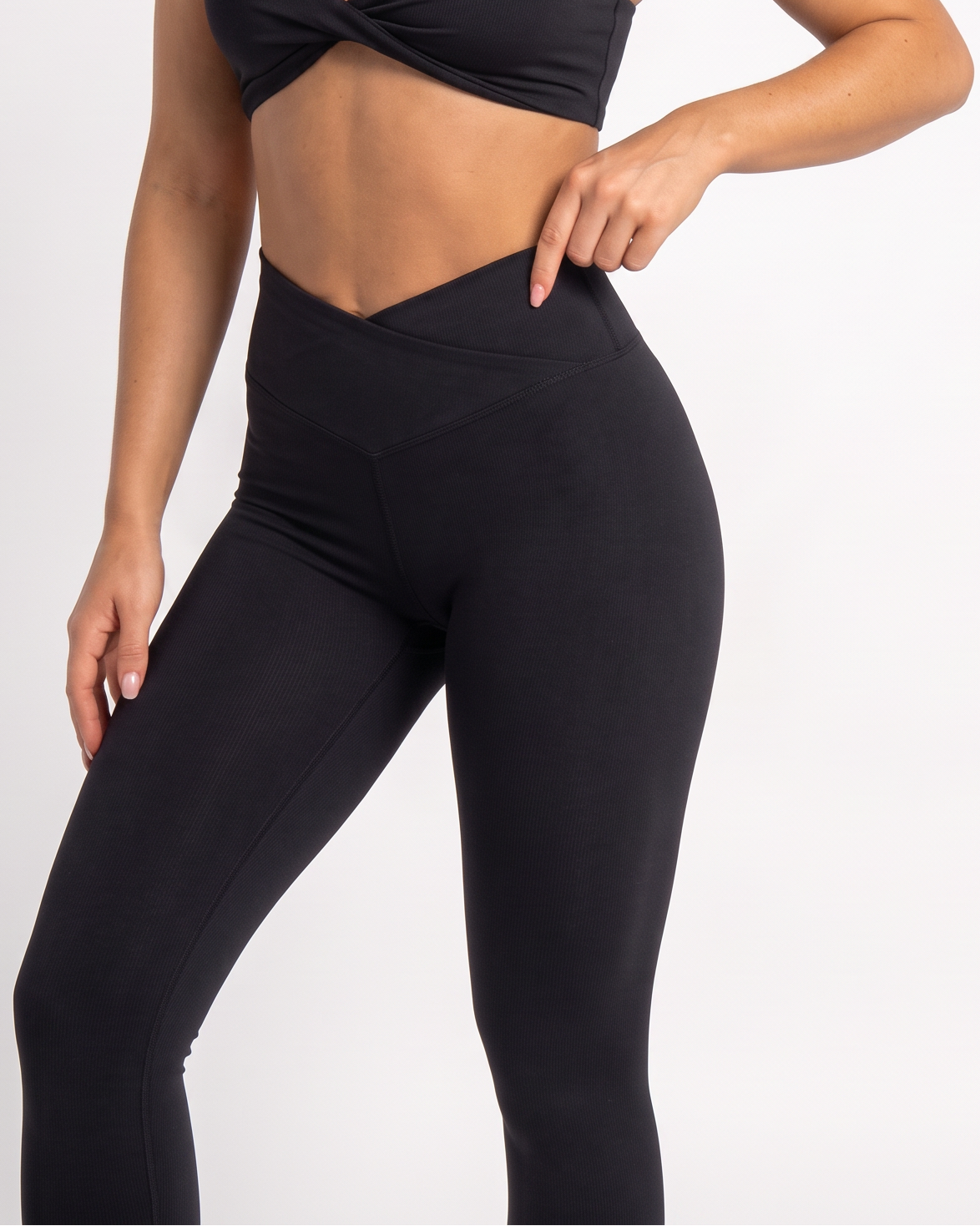 A woman modeling a black athletic set consisting of a crossover-front sports bra and high-waisted leggings, photographed against a clean light gray studio background.