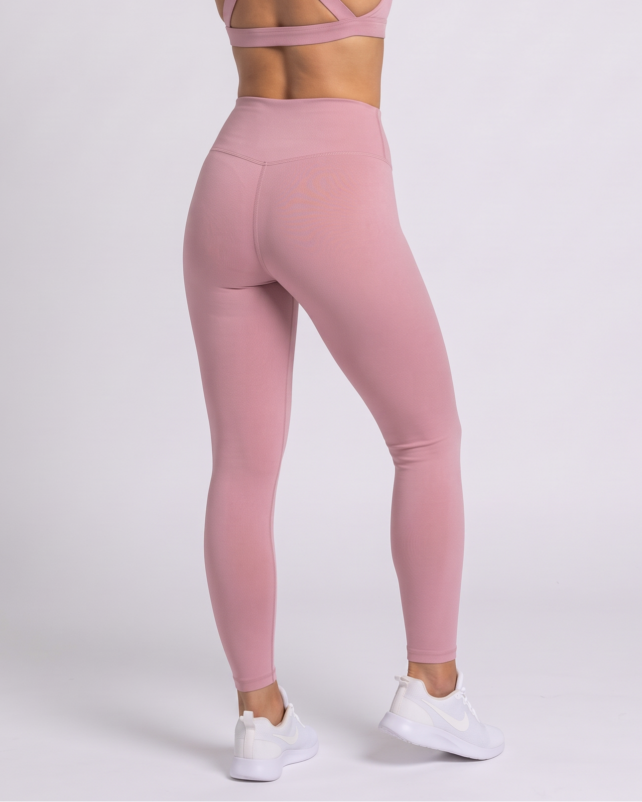 A woman shown from the back wearing a dusty pink sports bra and matching high-waisted leggings, paired with white sneakers, against a light gray studio background.