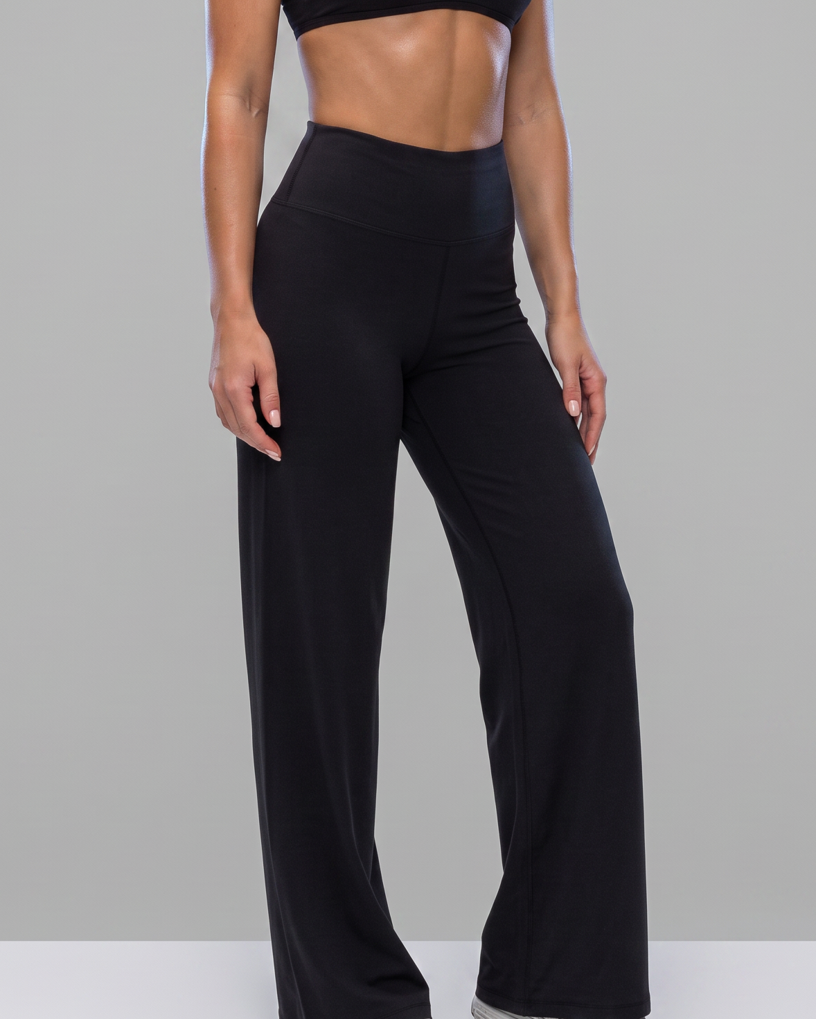 A woman modeling a black activewear set consisting of a sports bra and high-waisted wide-leg pants, standing against a solid gray studio background.