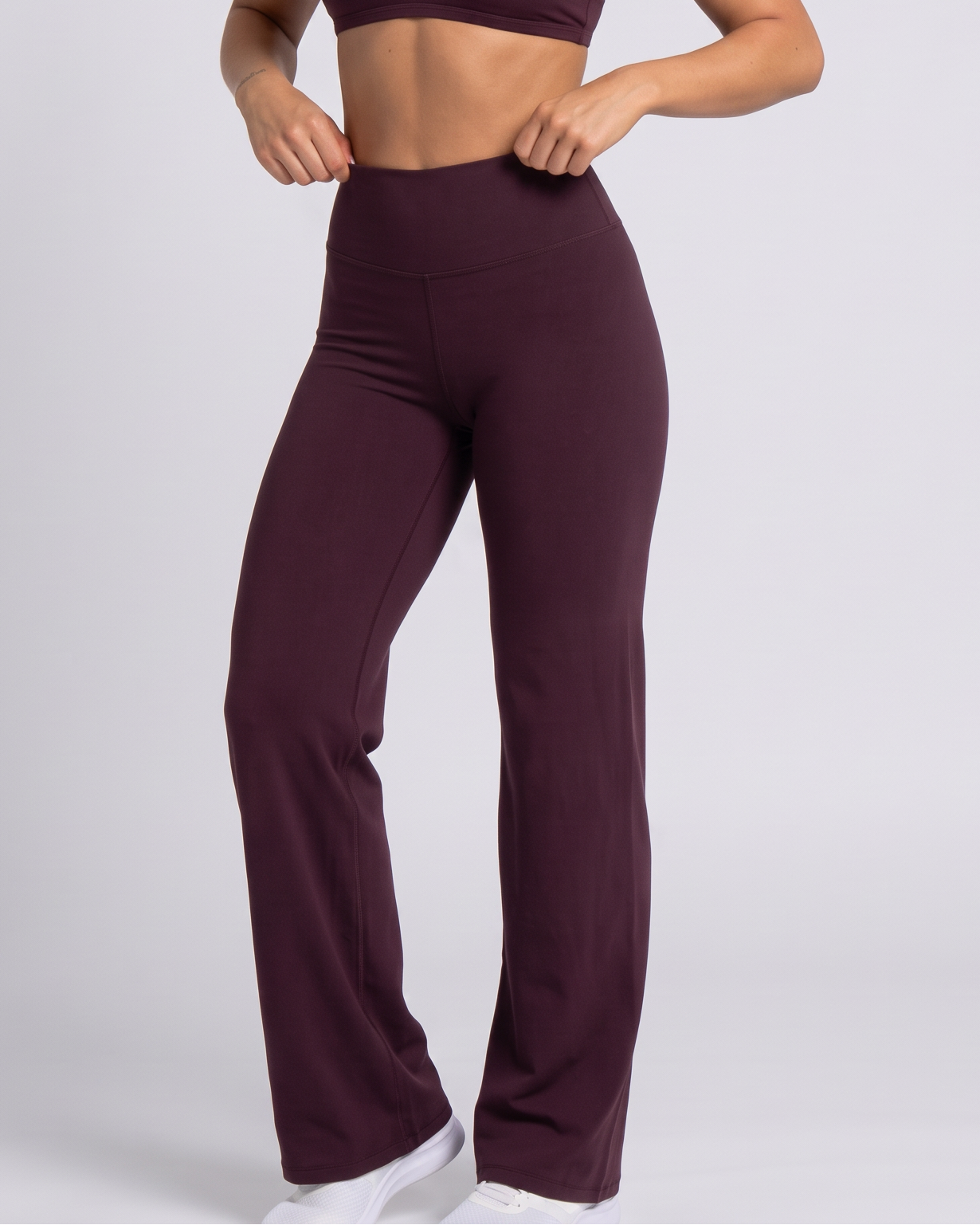 A woman modeling a matching athletic set consisting of a dark plum sports bra and high-waisted, wide-leg pants, paired with white sneakers, against a light gray studio background.