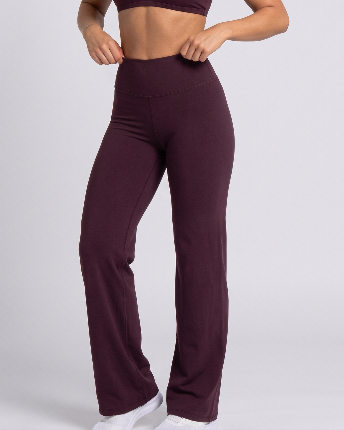 A woman modeling a matching athletic set consisting of a dark plum sports bra and high-waisted, wide-leg pants, paired with white sneakers, against a light gray studio background.