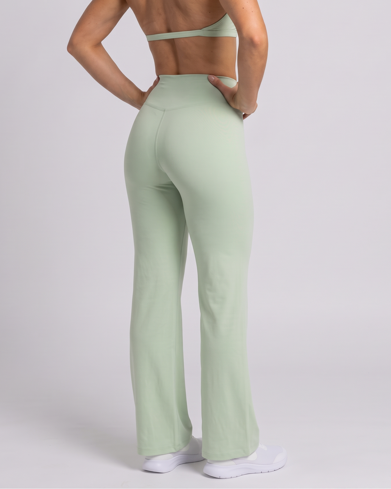 A woman with blonde hair in a ponytail is shown from the back wearing a sage green athletic set consisting of a halter crop top and matching high-waisted flared leggings, standing against a light gray studio background.