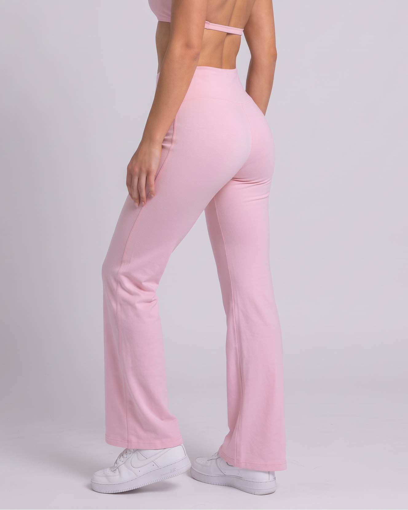 A woman modeling a light pink two-piece activewear set consisting of a halter crop top and matching high-waisted flared pants, paired with white sneakers against a light gray studio background.