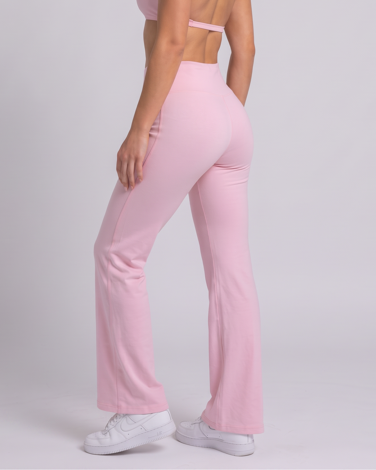 A woman modeling a light pink two-piece activewear set consisting of a halter crop top and matching high-waisted flared pants, paired with white sneakers against a light gray studio background.