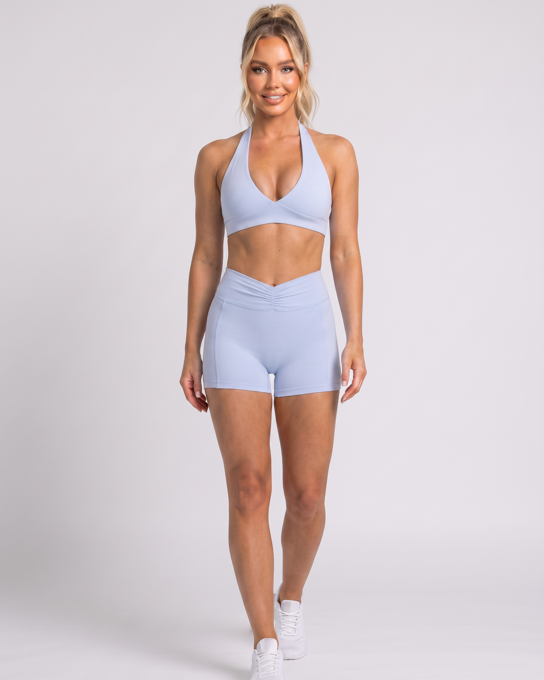 A woman modeling a light blue two-piece activewear set consisting of a halter-neck sports bra and matching high-waisted shorts, paired with white sneakers, against a light gray studio background.