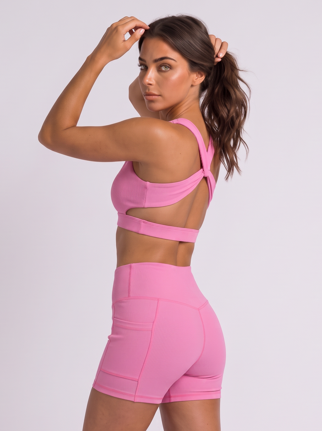 A woman modeling a bright pink athletic set consisting of a sports bra and matching high-waisted shorts, standing against a light gray studio background.