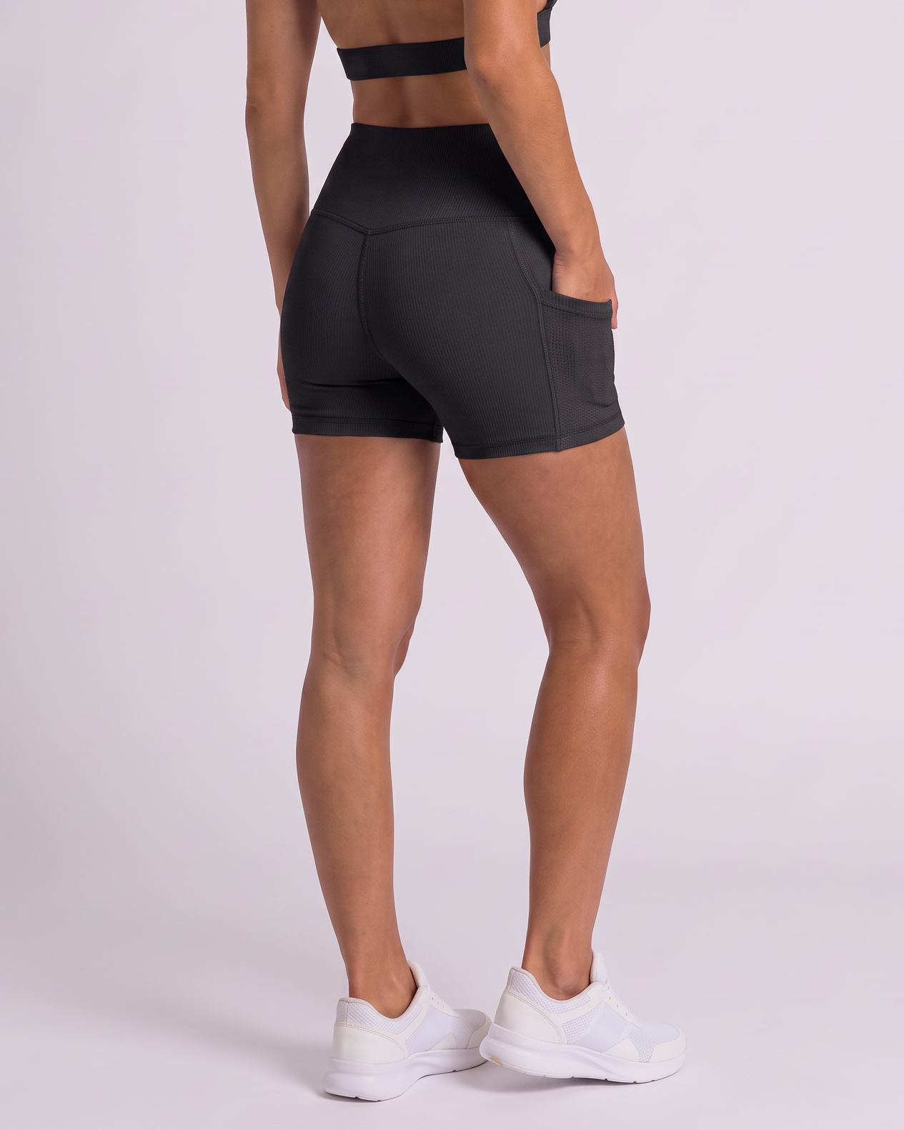 A woman modeling a black athletic set consisting of a sports bra with an open back and matching high-waisted shorts, shown from a rear three-quarter view against a light gray studio background.
