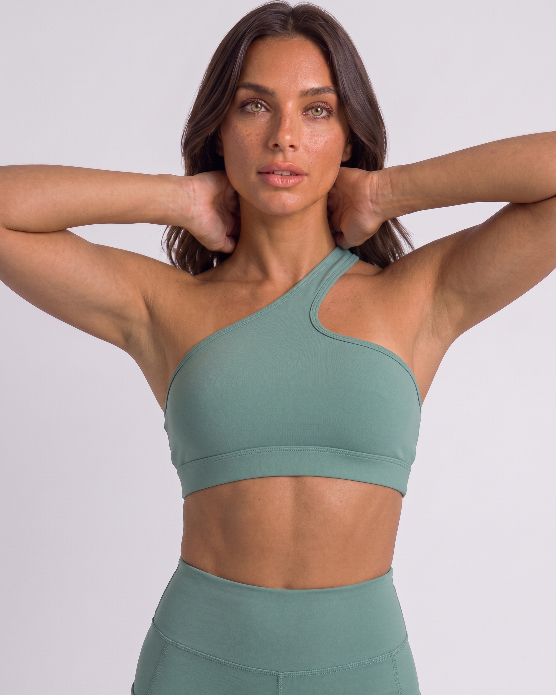 A woman modeling a sage green one-shoulder sports bra and matching high-waisted leggings against a light gray studio background.
