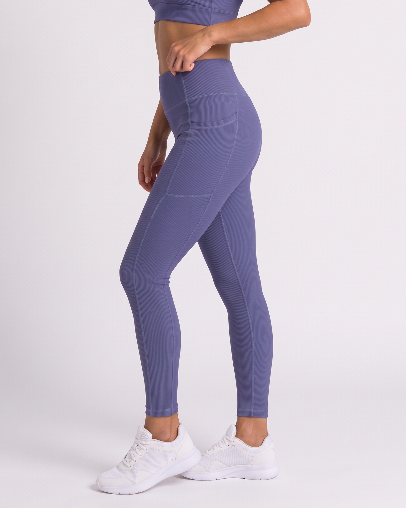A woman modeling a matching athletic set consisting of a dusty purple sports bra and high-waisted leggings, paired with white sneakers, against a plain light gray studio background.
