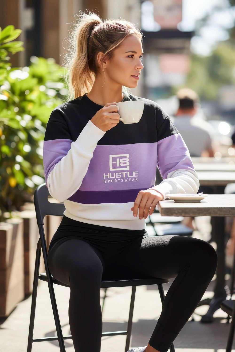 Woman wearing a black and purple 'Hustle Sportswear' sweatshirt sitting outdoors.