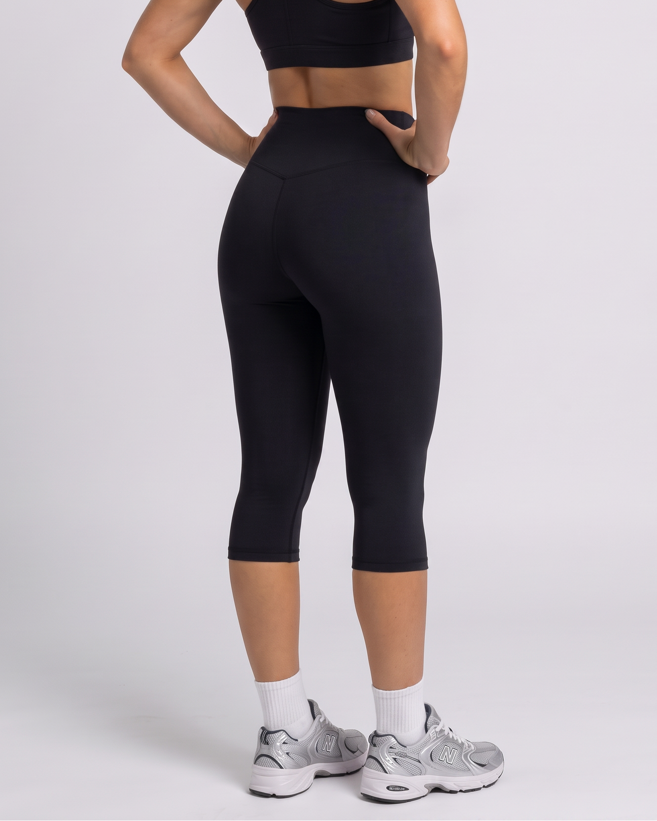A woman modeling a black sports bra and matching black high-waisted capri leggings, shown from a rear three-quarter angle against a light gray studio background.
