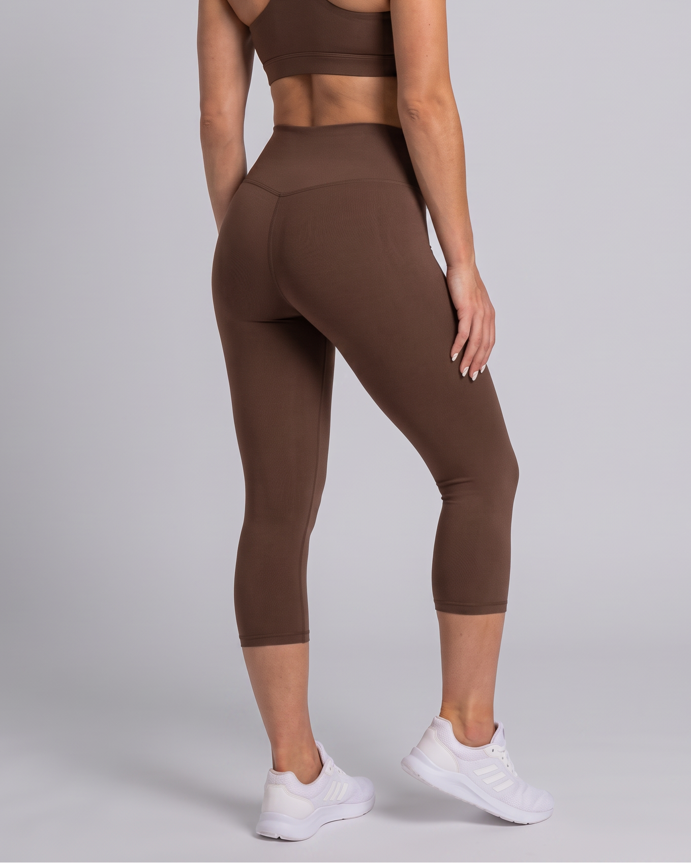 A woman modeling a matching brown activewear set, featuring a sports bra and high-waisted capri leggings, shown from a rear three-quarter angle against a light gray studio background.