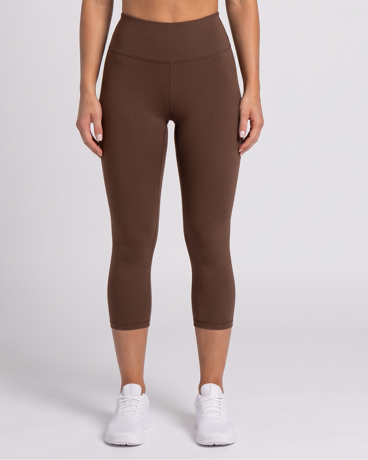 A woman modeling a matching brown activewear set consisting of a sports bra and high-waisted capri leggings, paired with white sneakers, against a light gray studio background.
