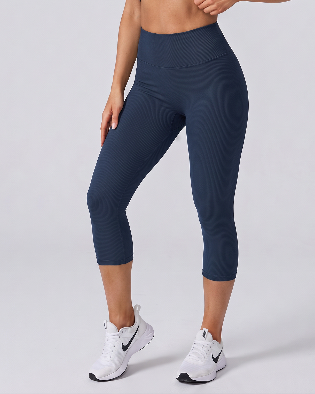 A woman modeling a matching navy blue activewear set consisting of a sports bra and high-waisted capri leggings, paired with white athletic sneakers, against a light gray studio background.